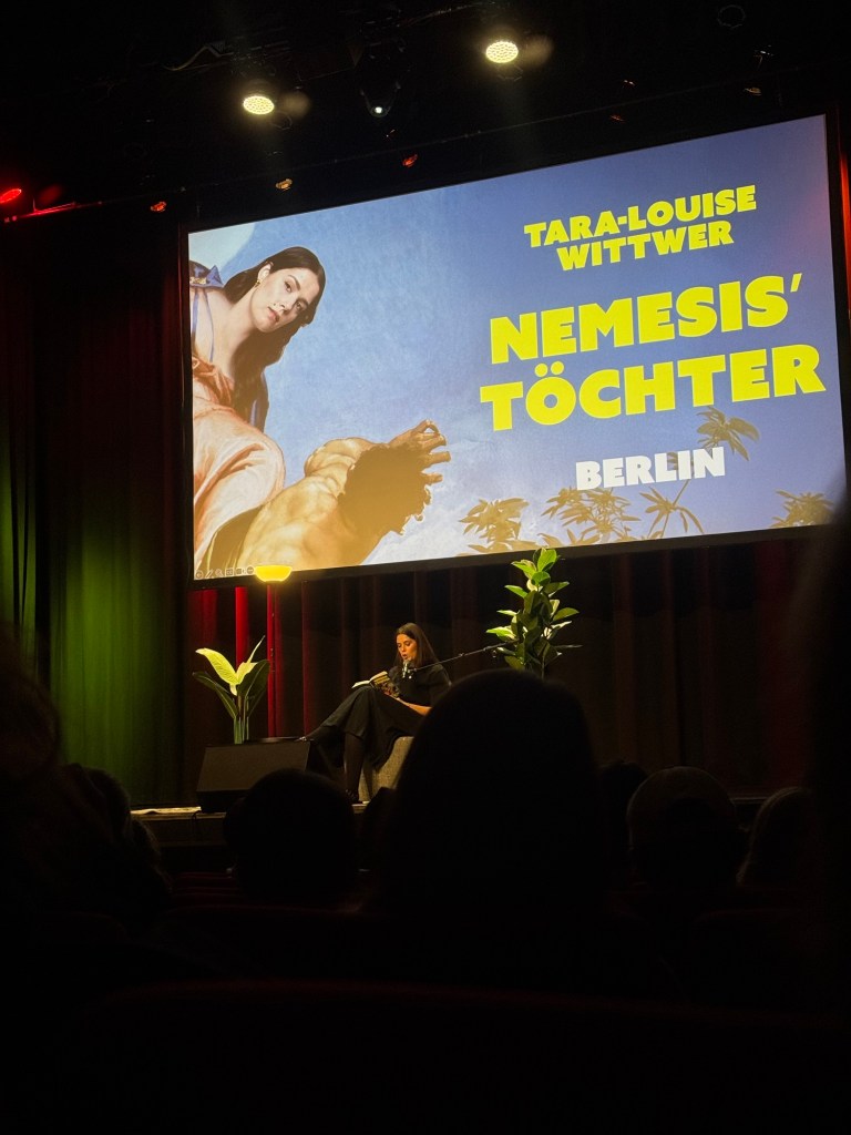 Bühne mit einer Großdarstellung des Covers von "Nemesis' Töchter" und Autorin Tara Wittwer bei der Lesung.